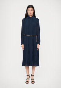 Γυναίκα ντυμένη με ένα ναυτικό μπλε πλισέ μίντι φόρεμα με μακριά διαφάνεια μανίκια, γιακά, χρυσή αλυσίδα μέση και μαύρα λουριά ψηλά τακούνια, standing σε λευκό φόντο.