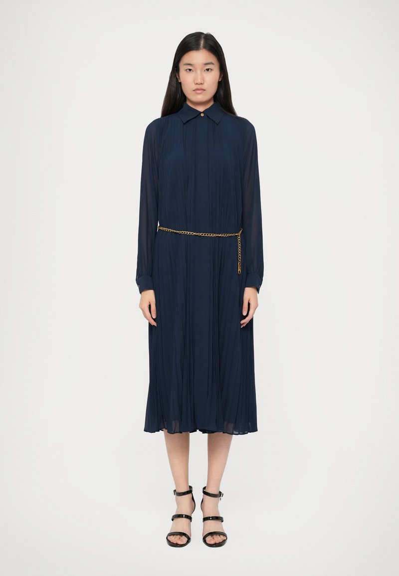 Γυναίκα ντυμένη με ένα ναυτικό μπλε πλισέ μίντι φόρεμα με μακριά διαφάνεια μανίκια, γιακά, χρυσή αλυσίδα μέση και μαύρα λουριά ψηλά τακούνια, standing σε λευκό φόντο.