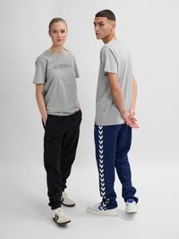 Twee personen staan frontaal en zijwaarts, dragen grijze Hummel T-shirts, zwarte en marineblauwe trainingsbroeken en witte sneakers tegen een effen achtergrond.