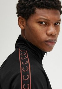 Giacca nera con zip e colletto alto, caratterizzata da strisce floreali color bordeaux lungo le maniche. Tessuto liscio e texturizzato.