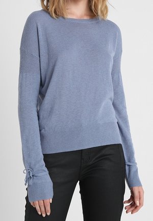 Mujer que lleva un suéter azul claro de manga larga con puños acanalados y lazos atados, combinado con pantalones negros, sobre un fondo liso.