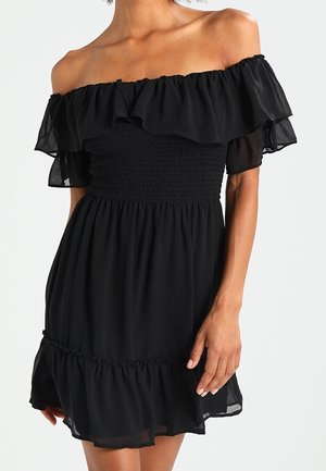 Woman wearing a black off-shoulder dress with smocked bodice and ruffled sleeves and hem, standing against a plain background.