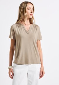 Krátký béžový top s výstřihem do V, nařasenými rameny a dekorativním prošíváním. Měkká tkanina, volný střih, kombinuje se s bílými širokými kalhotami.