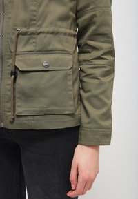 Olivově zelená bunda se zipem v přední �části, nastavitelným páskem v pase a dvěma předními kapsami, s kovovými prvky ve stříbrné barvě a hladkým povrchem.