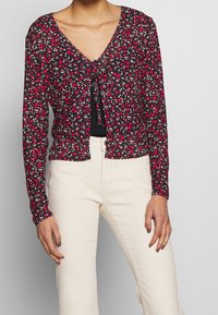 Mujer con una blusa negra de flores de manga larga atada en el pecho sobre una camiseta negra, combinada con pantalones blancos off-white de cintura alta.