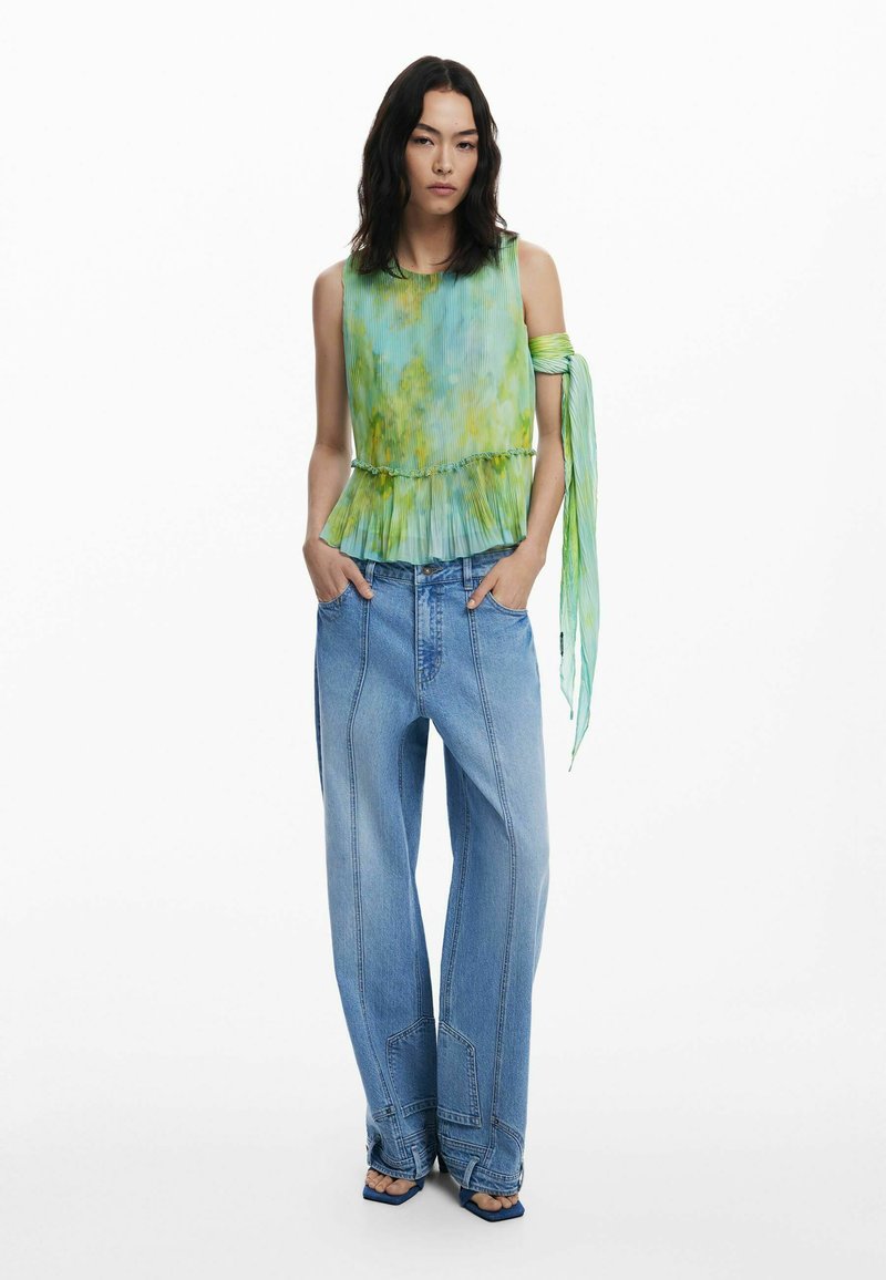 Ärmelloses Top in grün-gelbem Batikdesign mit Falten, kombiniert mit lässigen hellblauen Jeans mit auffälliger Naht und weit geschnittenen Beinen.