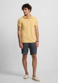 Camisa polo amarilla de manga corta con cuello, combinada con pantalones cortos grises. El modelo lleva zapatos de slip-on beige. Diseño minimalista, sin patrones visibles.