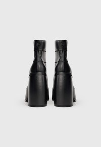 Botas de cano curto em pele preta com salto alto em bloco, design acolchoado e detalhes de costura visíveis. Bico arredondado e textura lisa.