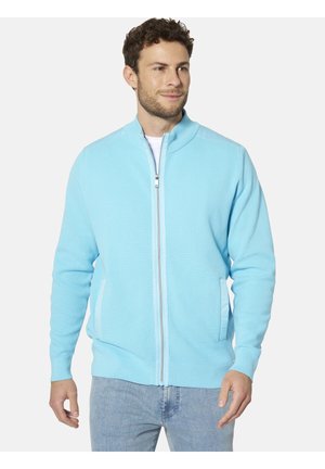 Mann trägt hellblaue, strukturierte Jacke mit Reißverschluss und hellblaue Jeans, steht und schaut leicht seitlich vor schlichtem Hintergrund.