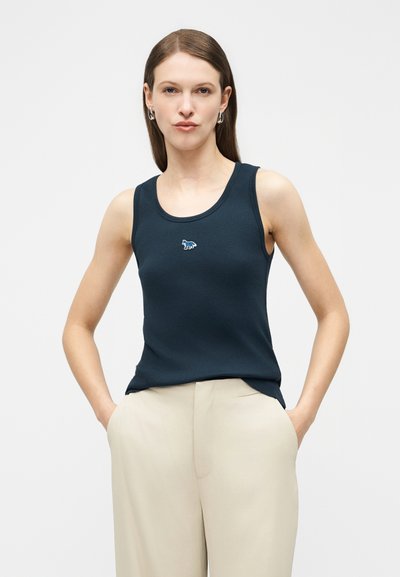 Γυναίκα με ίσια καστανά μαλλιά που φοράει ασημένια κρίκους σκουλαρίκια, σκούρο αμάνικο τοπ με μικρό λογότυπο και ανοιχτό μπεζ παντελόνι, με τα χέρια στις τσέπες.