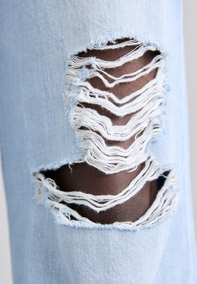 Ljusblå denimjeans med stora fransade hål som blottar ett mörkt lager underneath. Har fransade kanter och textil med struktur.