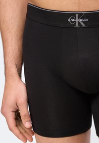 Mann trägt schwarze Calvin Klein Boxershorts, zeigt das Taillenband-Logo und die linke Hand mit kleinem Handgelenk-Tattoo.