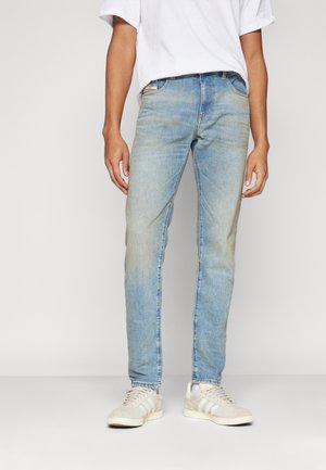 Personne portant un jean slim bleu clair, des baskets blanches et une chemise blanche à manches courtes, debout devant un fond uni.