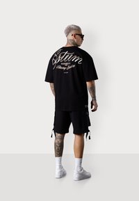 Schwarzes Oversized-T-Shirt mit weißem Schriftzug auf dem Rücken, kombiniert mit schwarzen Cargo-Shorts, weißen Sneakern und weißen Socken.