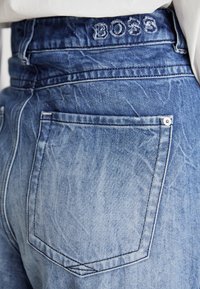 Μπλε τζιν παντελόνι με ψηλή μέση, πίσω τσέπη και το όνομα της μάρκας "BOSS" κεντημένο στη ζώνη σε αντιθετικό λευκό νήμα.