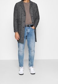 Mann in einem grauen Mantel mit Fischgrätmuster, braunem Hemd, hellblauen Jeans, schwarzem Gürtel und weißen Nike-Sneakern, der mit einer Hand in der Tasche steht.