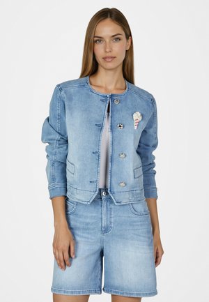 Young woman wearing light blue denim jacket with silver buttons and matching knee-length shorts, standing against white background.