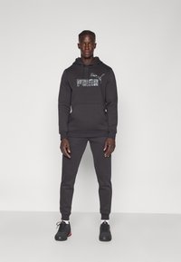 Felpa nera con logo "PUMA" grigio e tasca a marsupio, abbinata a pantaloni della tuta coordinati. Completo realizzato in tessuto morbido, con polsini a coste.