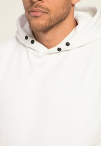 Vit hoodie med en mjuk textur, med en stor huva och tre svarta tryckknappar vid halsen för stängning och detalj.