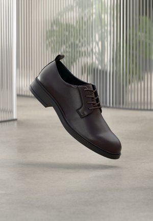 Chaussure en cuir marron foncé avec une texture lisse, design à lacets, bout arrondi, et une semelle basse en caoutchouc noir. Agrémentée d'une bride arrière pour faciliter l'enfilage.