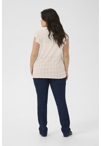 Haut beige clair avec des détails en œillets, manches volantées, associé à un jean noir à pattes d'eph, vue de dos. Texture lisse, style décontracté.