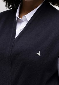 Chaleco de lana azul marino con diseño de cuello en V, que presenta un pequeño logo plateado en el pecho. La textura parece suave y de punto fino.