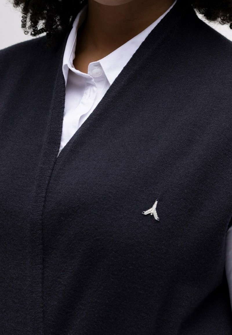 Chaleco de lana azul marino con diseño de cuello en V, que presenta un pequeño logo plateado en el pecho. La textura parece suave y de punto fino.