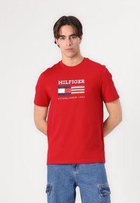 Červené bavlněné tričko s kulatým výstřihem a krátkými rukávy. Obsahuje bíle, modře a červeně tištěný text "HILFIGER" a design vlajky.