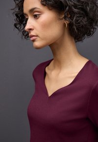 Burgunderfarbenes V-Ausschnitt-Oberteil aus glattem Stoff, mit taillierter Silhouette und kurzen Ärmeln, das ein sauberes, modernes Design betont.