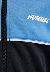 Niebieska i czarna sportowa kurtka z zamkiem, z białymi akcentami i logo "HUMMEL" w białym kolorze. Gładka tekstura, sportowy design.