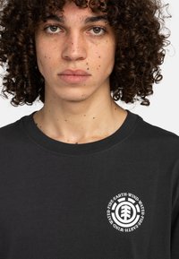 Zwarte kortemou t-shirt van katoen, met een ronde witte logo op de linkerborst en tekst die de elementen beschrijft: aarde, lucht, vuur, water.