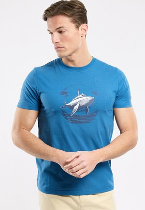 Homme portant un t-shirt bleu avec un graphisme de baleine, regardant vers sa gauche, les mains jointes, debout devant un fond blanc.