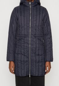 Mujer con una larga chaqueta acolchada azul marino, con cremallera frontal, capucha y bolsillos laterales, de pie contra un fondo claro y liso.