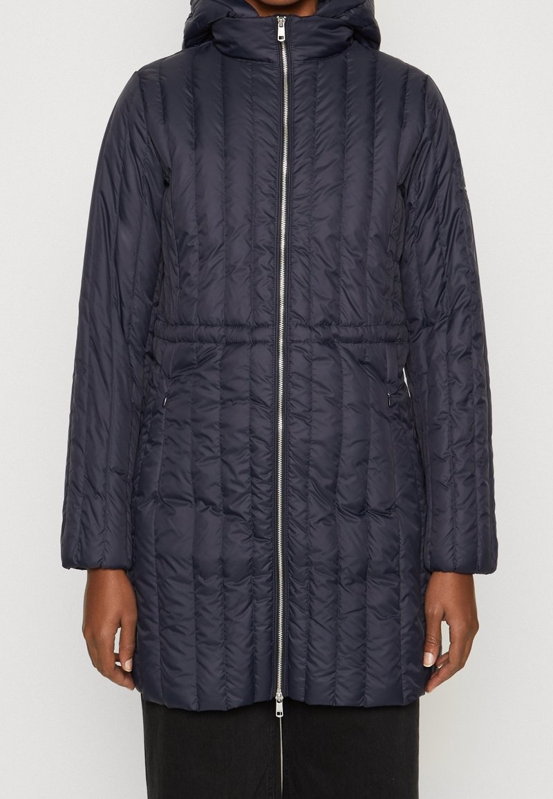Mujer con una larga chaqueta acolchada azul marino, con cremallera frontal, capucha y bolsillos laterales, de pie contra un fondo claro y liso.