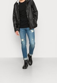 Chaqueta impermeable negra con capucha, camiseta negra, vaqueros azules desgastados y botines negros con puños enrollados y costuras en contraste.