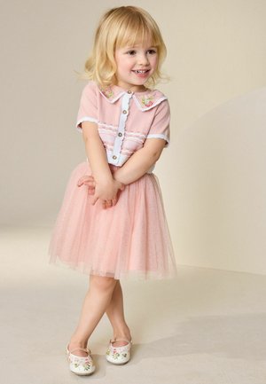 Lächendes junges Mädchen mit blondem Haar, das ein pinkfarbenes Oberteil mit Blumenkrause und einen glitzernden pinkfarbenen Tüllrock trägt, steht mit überschlagenen Beinen.