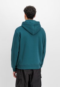 Sudadera con capucha en color azul verdoso, confeccionada en un tejido suave, con un bolsillo tipo canguro y puños acanalados. El diseño es de corte holgado y cuenta con una capucha sin costuras.