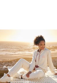 Felpa e pantaloni della tuta color crema con una grafica solare, cordoncino con accenti colorati, abbinati a sneaker bianche. Abbigliamento casual e accogliente.