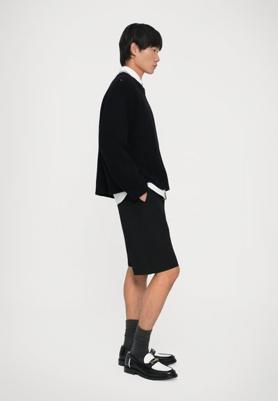 Ung mand iført sort strikket sweater, hvid skjorte, sorte shorts, grå sokker og sort-hvide loafers, der står i profil mod en hvid baggrund.