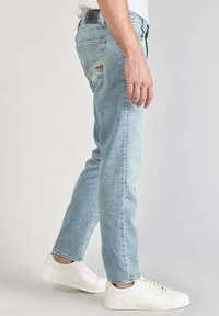 Jeans de mezclilla azul claro con corte slim, que presentan detalles desgastados, cinco bolsillos y un parche de piel en la cinturilla trasera.