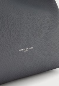 Sac à main en cuir gris foncé avec une surface texturée. Présente un petit logo de marque en blanc, mettant en avant un design minimaliste.