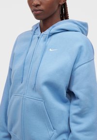 Άτομο που φοράει μια ανοιχτόγαλανη ζακέτα Nike με φερμουάρ, εμπρόσθια τσέπη και κουκούλα με κορδόνι, πάνω σε απλό φόντο.