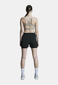 Sportbeha in beige met een kruisrugdesign, gecombineerd met zwarte sportshorts. Het model draagt witte sokken en lichtgrijze hardloopschoenen.