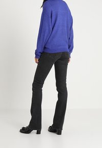 Personne portant un pull à manches longues bleu, un jean noir bootcut ajusté, et des chaussures noires à talons avec des pampilles, debout, vue de dos.