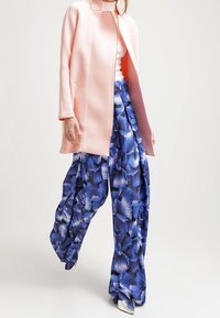 Abrigo largo de color rosa claro con un cuello alto sobre una blusa blanca, combinado con pantalones de pierna ancha de estampado floral azul y zapatos plateados.