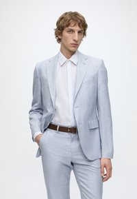 Jeune homme portant un costume gris clair et une chemise blanche, debout avec une main dans la poche devant un fond blanc uni.