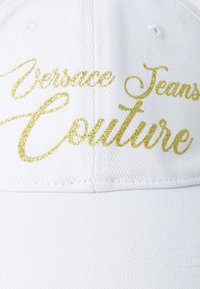 White cap made of textured fabric, featuring gold glittery text “Versace Jeans Couture” prominently on the front.