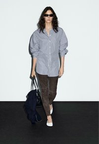 Camicia a righe con maniche a sbuffo, pantaloni scamosciati marrone scuro, scarpe bianche flat e borsa nera, su uno sfondo semplice.