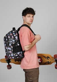 Mochila negra con patrones de dibujos animados coloridos, múltiples compartimentos con cremallera, llevada en un hombro. Patineta de madera con ruedas rojas exhibida.