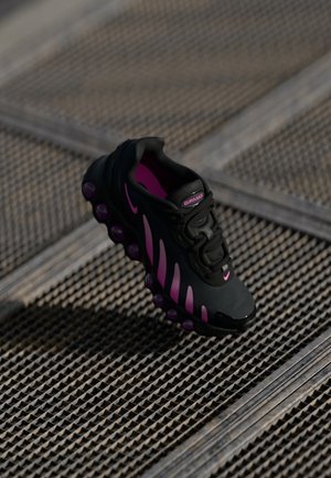 Czarny i fioletowy but sportowy Nike Air Max zawieszony nad teksturowaną metalową kratą podłogi, podkreślający podeszwę i boczny design.